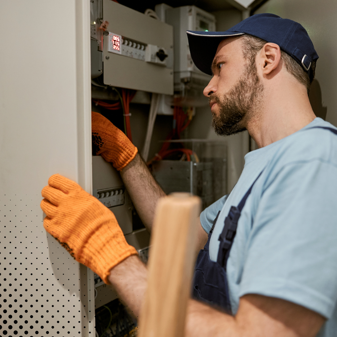 A home inspector wearing orange gloves and a blue cap carefully examines an electrical panel.
